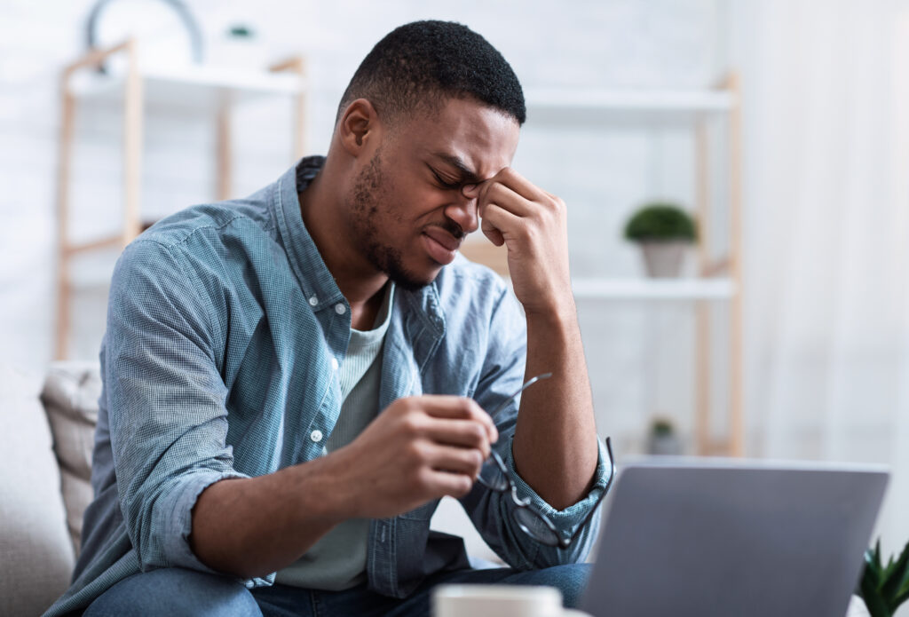 Eyes Fatigue. African Man Massaging Nosebridge Tired After Work On Laptop Computer Sitting On Couch Indoors. Headache And Eye Sight Problem Symptoms, Eyestrain, Glaucoma Concept